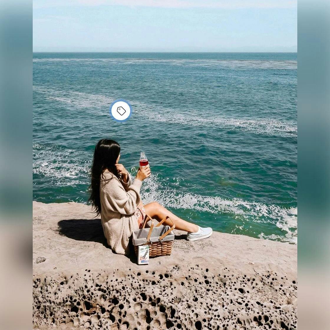 A woman is sitting on a cliff in front of water. She is drinking True Lemon Energy.