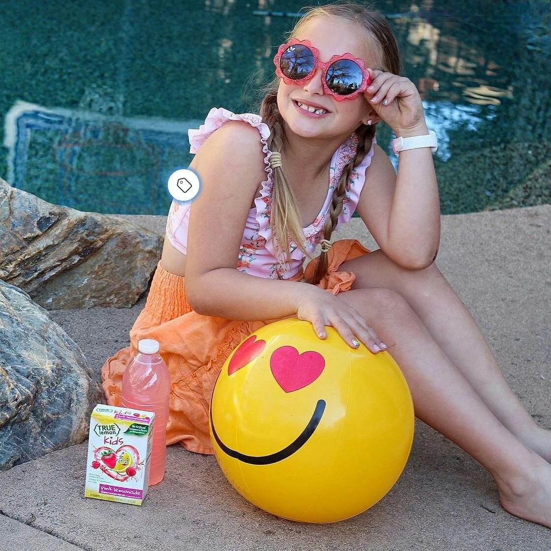 A girl is sitting by the pool with a beach ball and some True Lemon Kids.