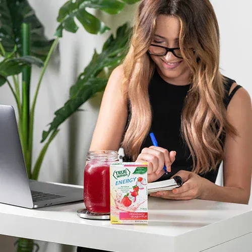 A woman is sitting at a desk writing in a notebook. On the table is a box of True Lemon Energy Strawberry Dragonfruit.