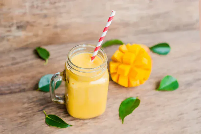 A mango smoothie in a glass on a table with a sliced mango behind it.