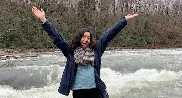 Mia is standing in front of a river. She is smiling and has both arms outstretched over her head.